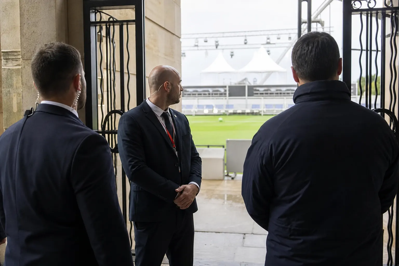 des agents de sécurité devant les grilles d'un stade, pelouse verte au loin